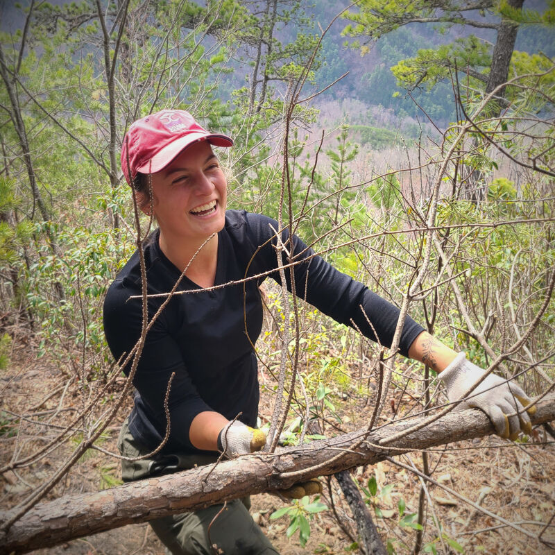 A young woman with a red cap is smiling and working outdoors. She is wearing gloves and holding a branch, possibly clearing a trail in a wooded area. The background shows a scenic view with trees and mountains, suggesting she is in a natural environment. Her attire includes a long-sleeved black shirt and khaki pants, suitable for outdoor work.